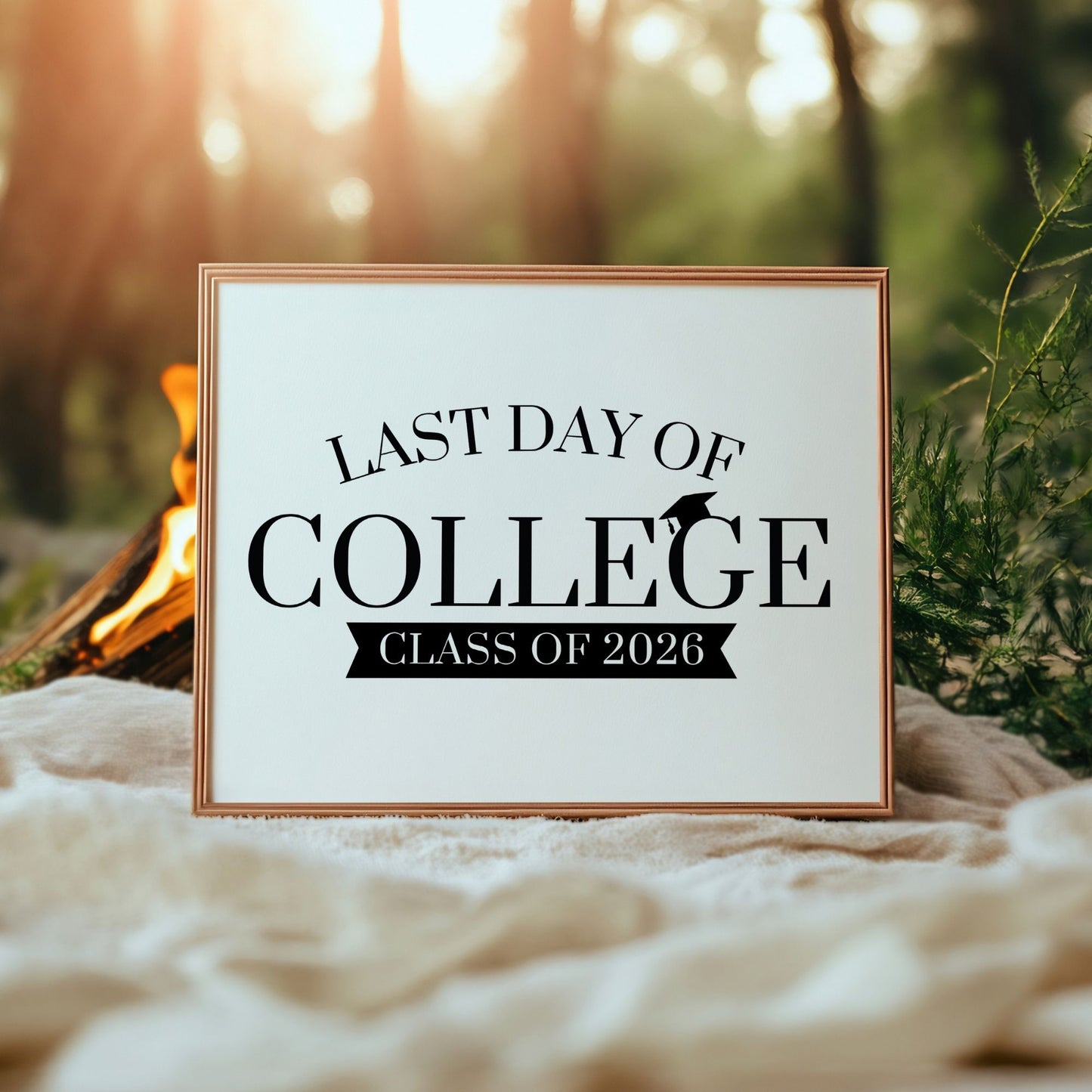 Framed sign with 'Last Day of College Class of 2026' text in a natural setting with a campfire and trees.