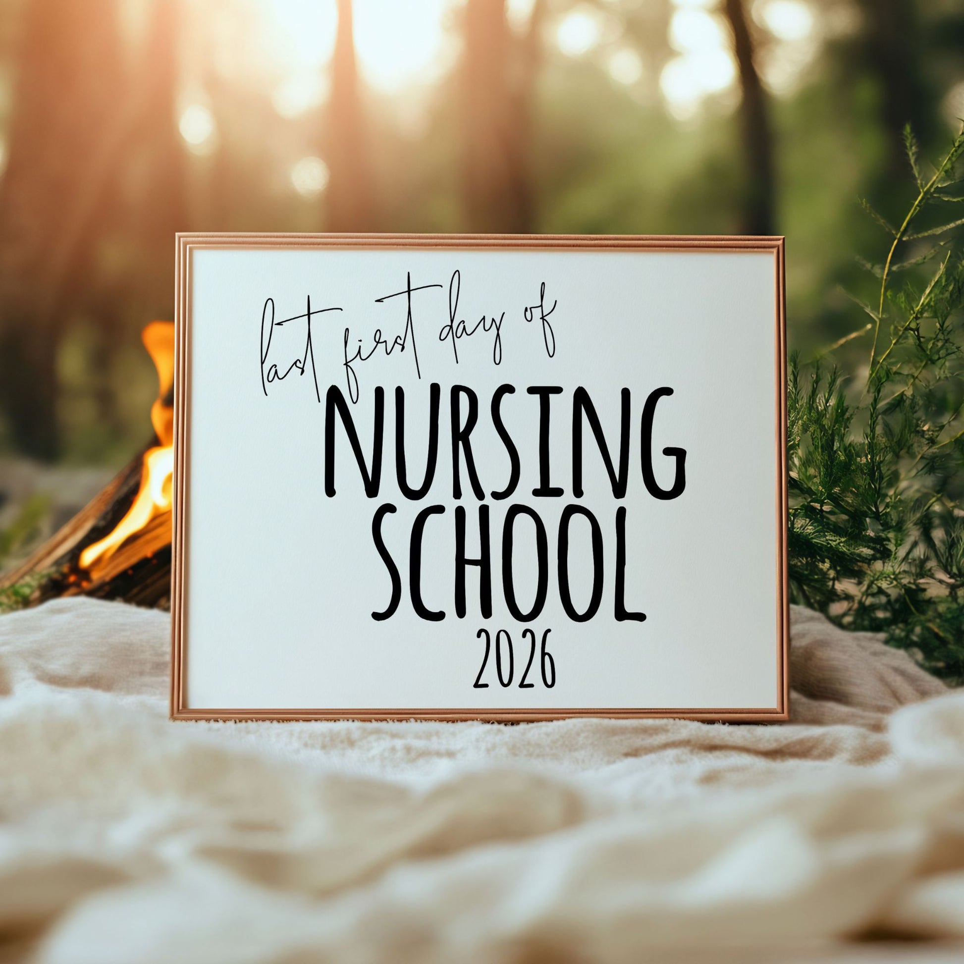 A landscape-oriented nursing school sign resting on a soft white blanket outdoors, framed by warm golden hour sunlight and greenery. The sign reads 'Last First Day of Nursing School 2026' in modern black typography