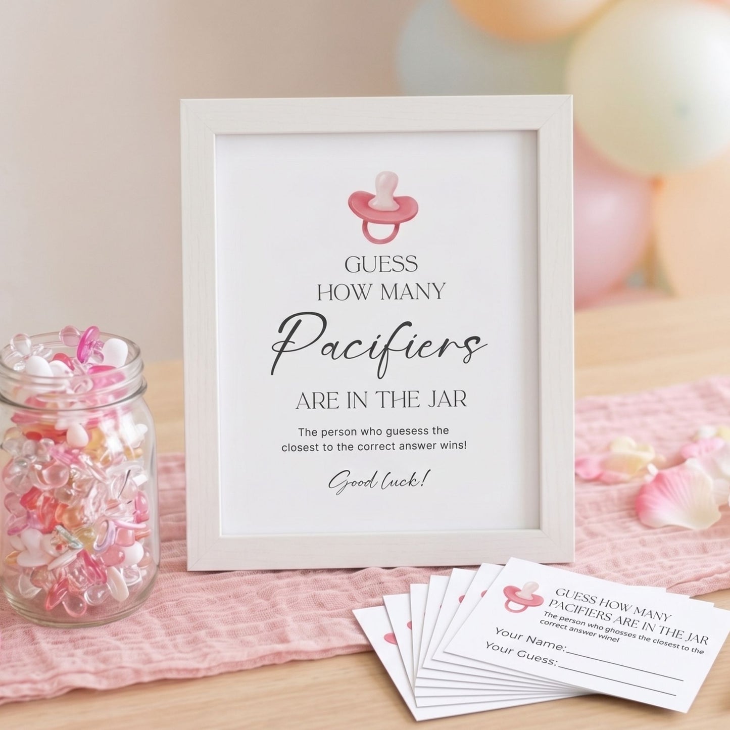 A baby shower game sign in a white frame that reads 'GUESS HOW MANY Paciers ARE IN THE JAR' with a pink pacifier icon, displayed on a pink cloth with matching cards and a jar of pastel candies.