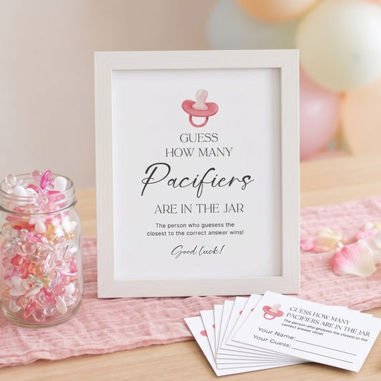 A baby shower game sign in a white frame that reads 'GUESS HOW MANY Paciers ARE IN THE JAR' with a pink pacifier icon, displayed on a pink cloth with matching cards and a jar of pastel candies.