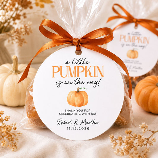A round white gift tag with an orange ribbon bow, wrapped around a clear cellophane bag containing a pumpkin-themed treat. The tag reads 'a little PUMPKIN is on the way!' with a small pumpkin illustration and personalized text for Robert & Martha.
