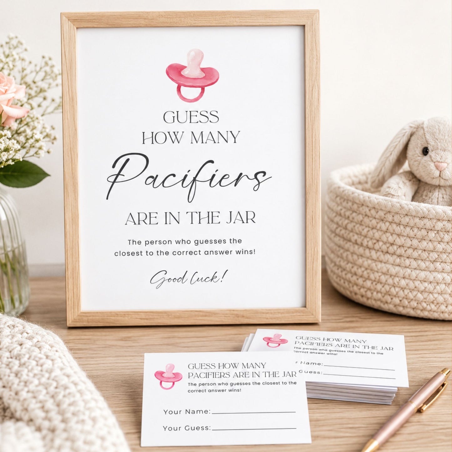A framed baby shower sign reading 'Guess how many pacifiers are in the jar' with a pink pacifier icon, displayed on a wooden surface with matching printed cards, a stuffed bunny in a basket, flowers, and a knit blanket.