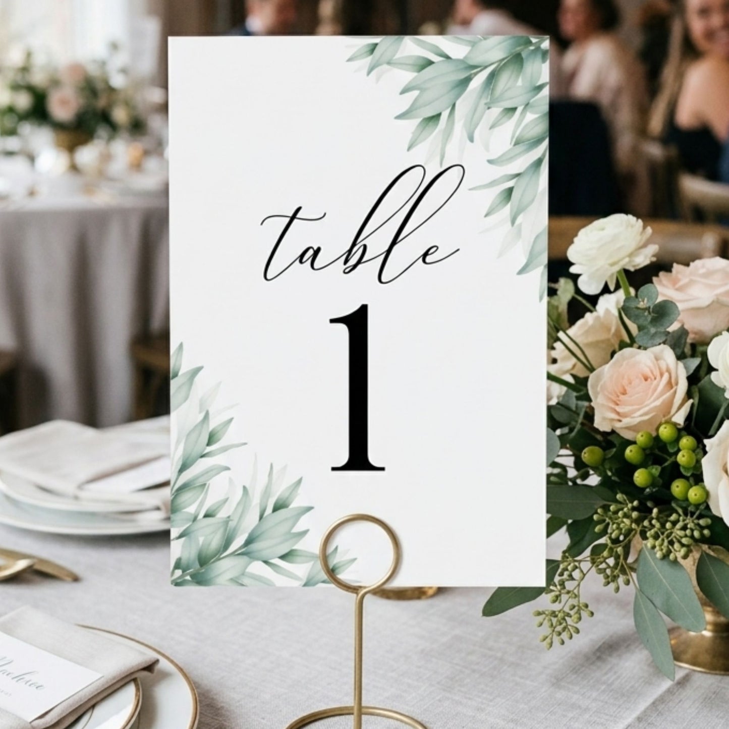 Table number card with greenery design on a table setting with flowers and plates.