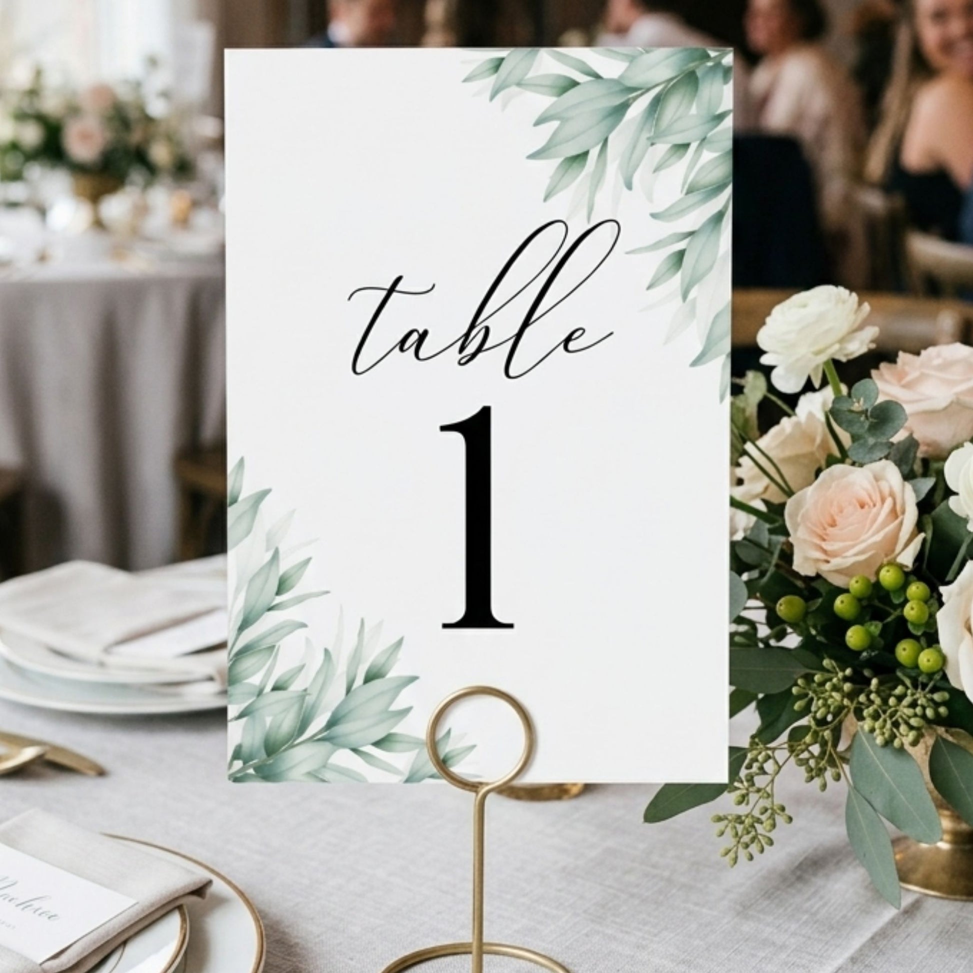 Table number card with greenery design on a table setting with flowers and plates.