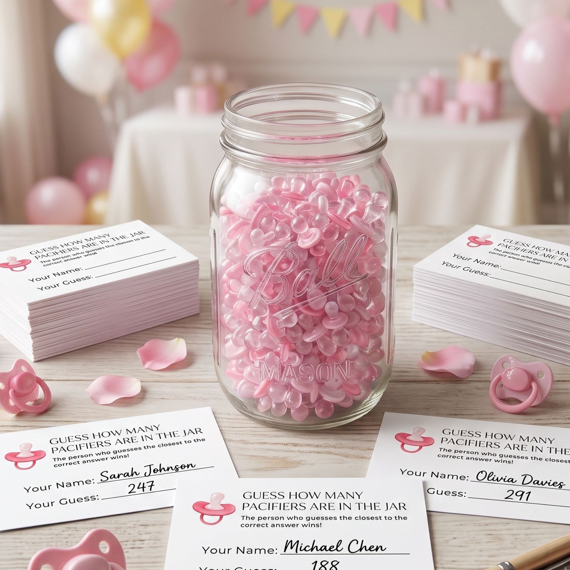 A clear glass mason jar filled with pink pacifiers sits on a tabletop, surrounded by printed guest name cards and scattered pink pacifiers in a softly decorated party setting.