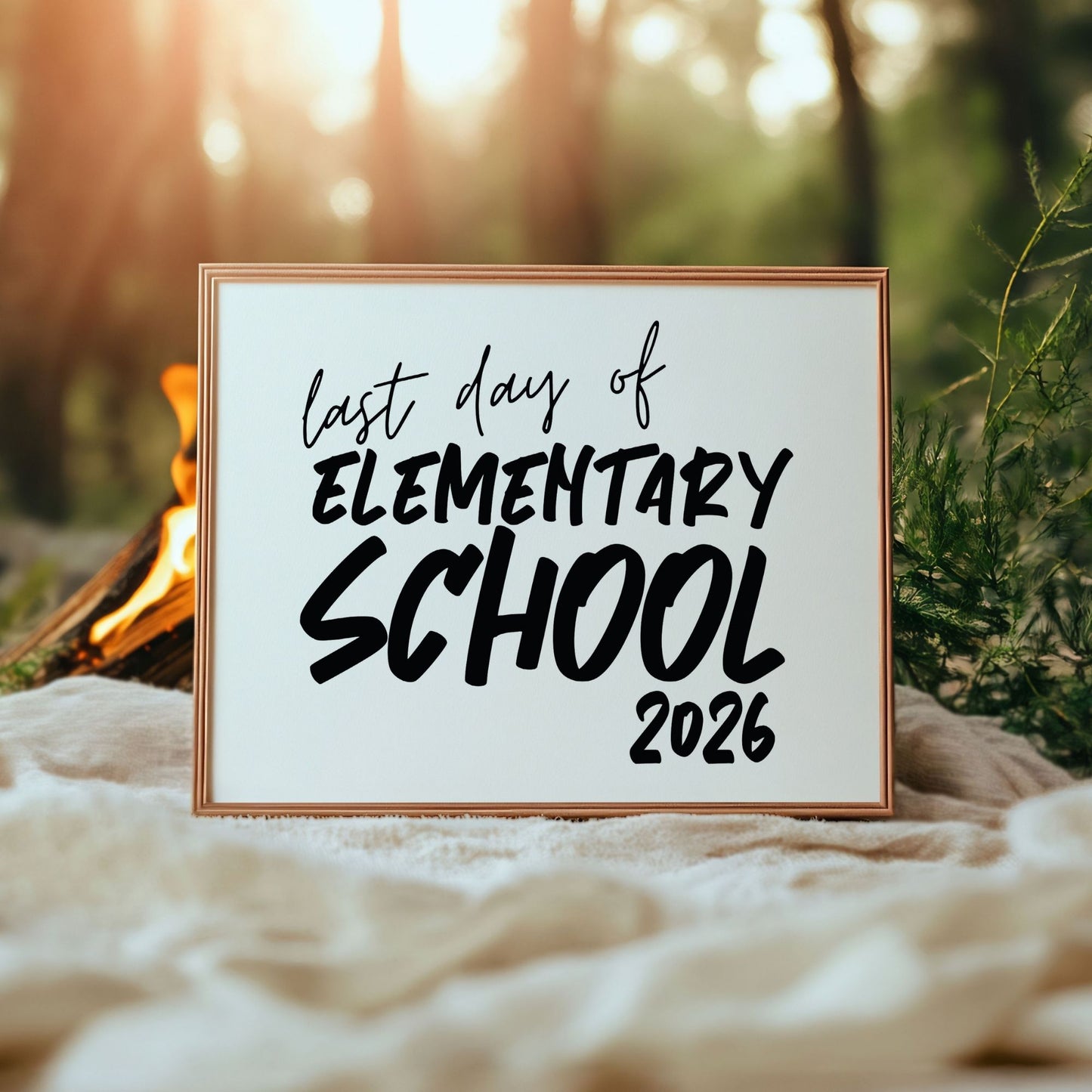Framed sign with 'last day of Elementary School 2026' text in a natural setting with a campfire and trees.