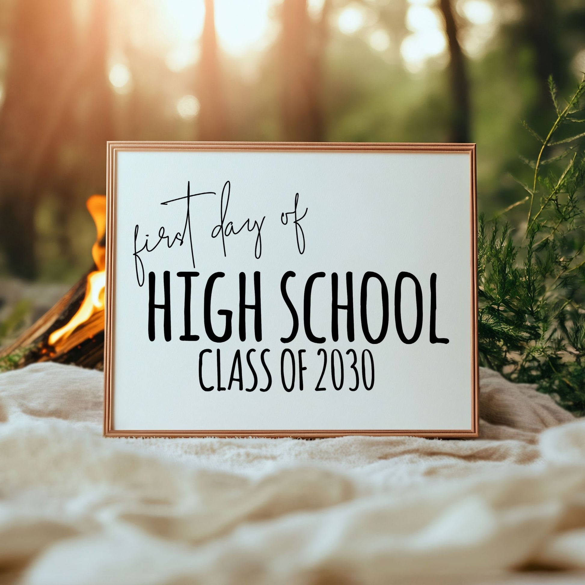 Framed sign with 'first day of High School Class of 2030' text in a natural setting with a campfire.