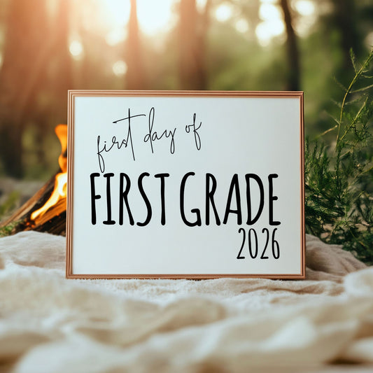 Framed sign with 'first day of first grade 2026' text in a natural setting with a campfire.