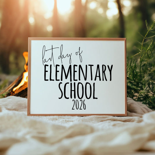 Framed sign with 'first day of Elementary School 2026' text in a natural setting with a campfire.