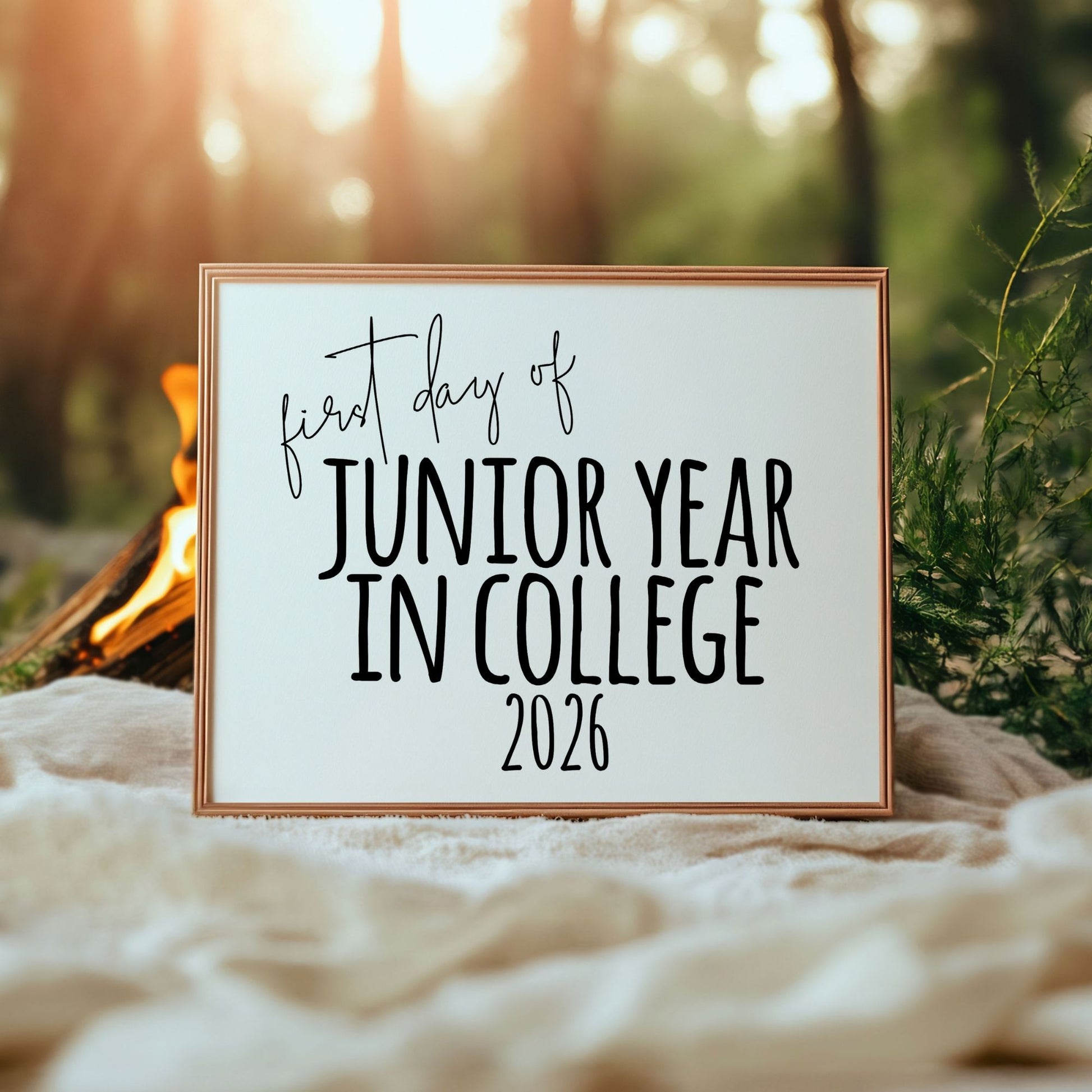 Sign with 'first day of Junior Year in College 2026' text in a forest setting with a campfire.