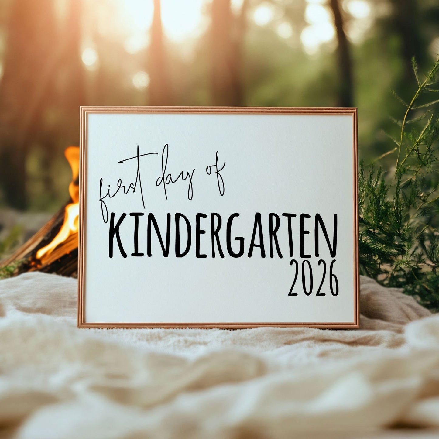 Framed sign with 'first day of Kindergarten 2026' text in a natural setting with a campfire and trees.