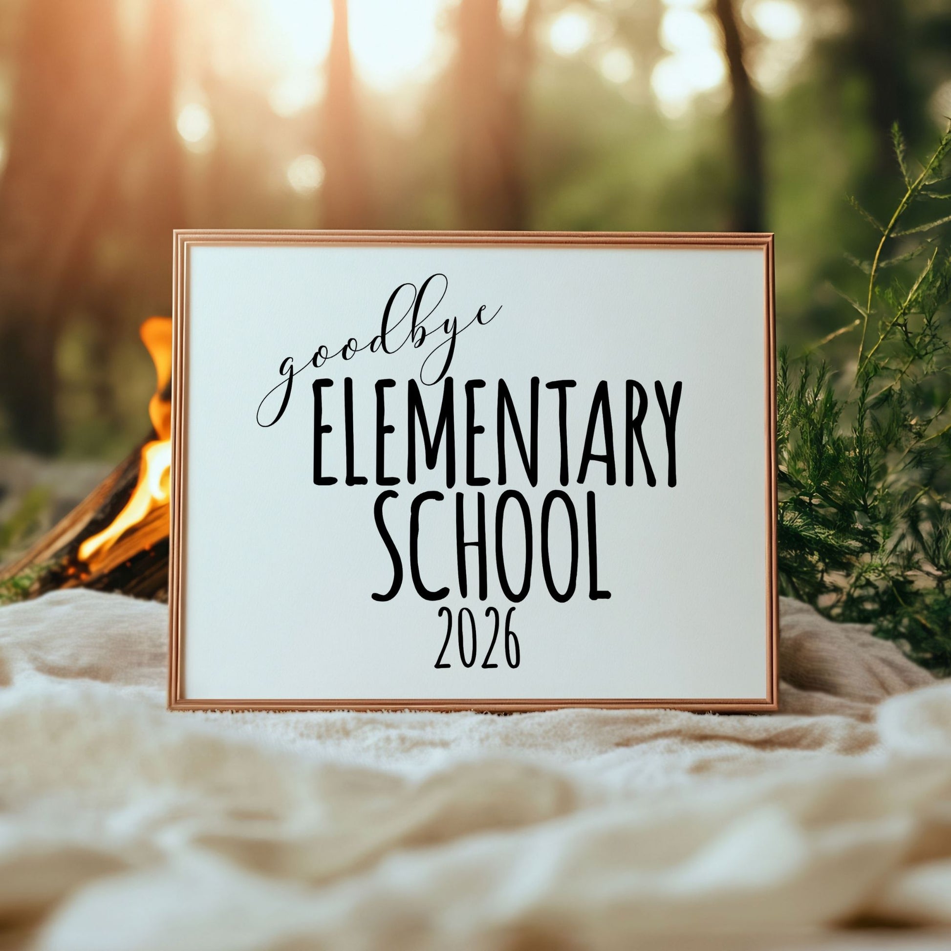 Framed sign with 'goodbye Elementary School 2026' text in a natural setting with a campfire.