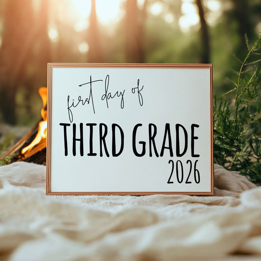 Framed sign with 'first day of third grade 2026' text in a natural setting with a campfire.