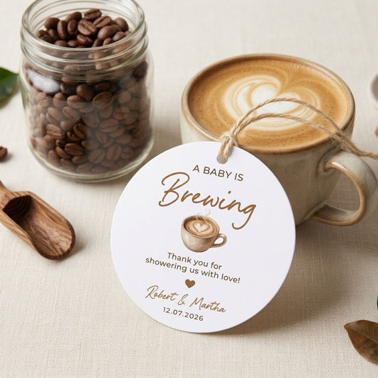 A personalized round white coffee-themed shower favor tag with a rustic twine string, displayed next to a cup of latte art coffee and a glass jar of coffee beans on a beige fabric surface.