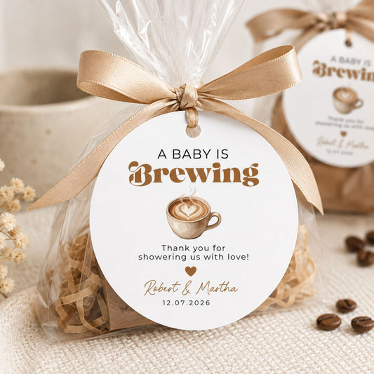 A clear cellophane gift bag tied with a beige ribbon and a round white tag that reads 'A BABY IS Brewing' with a coffee cup illustration and personalized text, styled on a neutral fabric surface with coffee beans and a blurred mug in the background.