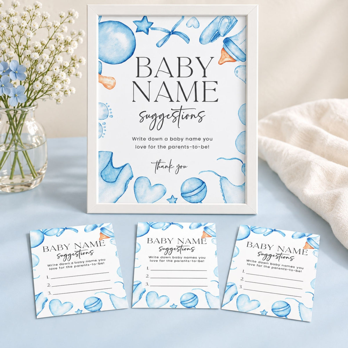 A framed baby name suggestion print with blue watercolor illustrations of balloons, hearts, stars, and pacifiers, shown alongside three matching printed cards on a light blue surface with a white towel and flowers in a vase.