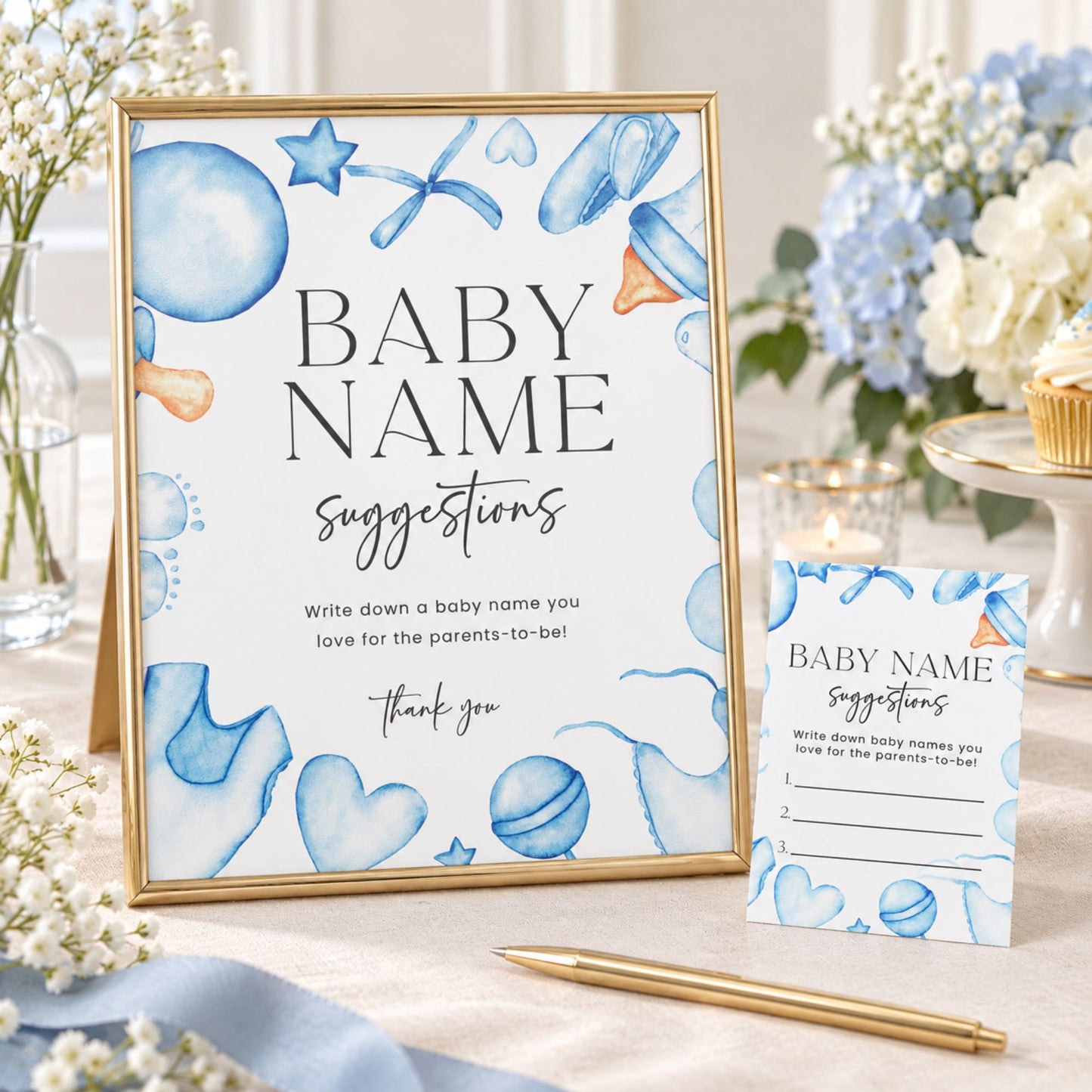 A gold-framed baby name suggestion sign and matching card stand on a tabletop with a pen nearby. The sign and card have a white background with blue watercolor illustrations of balloons, hearts, stars, and baby items, plus black text reading 'BABY NAME suggestions' and a thank you message.