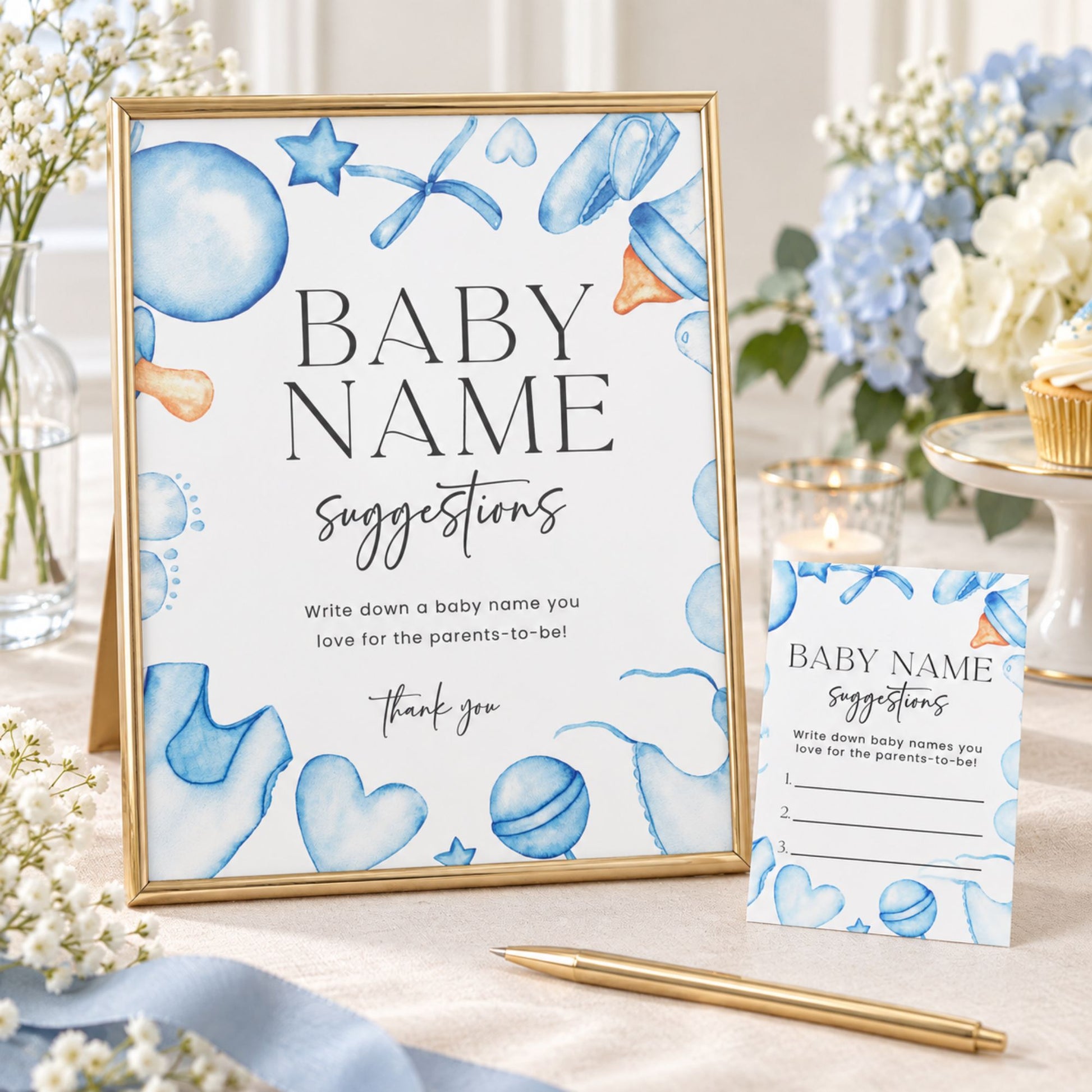 A gold-framed baby name suggestion sign and matching card stand on a tabletop with a pen nearby. The sign and card have a white background with blue watercolor illustrations of balloons, hearts, stars, and baby items, plus black text reading 'BABY NAME suggestions' and a thank you message.