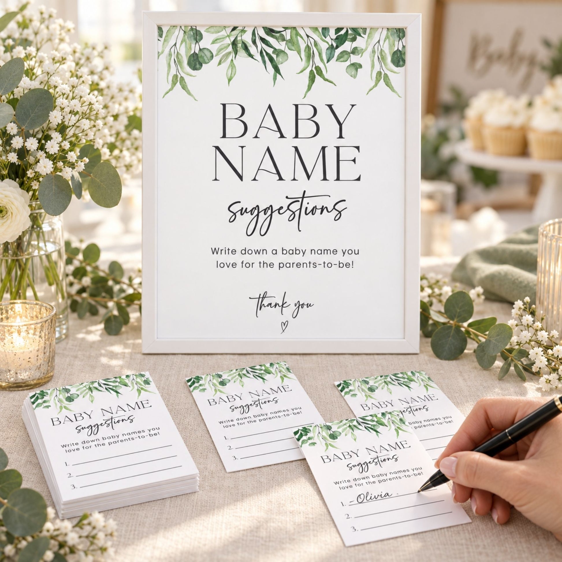 A baby shower sign and matching cards with green leaf illustrations, displayed on a table with flowers, candles, and a hand writing on a card.