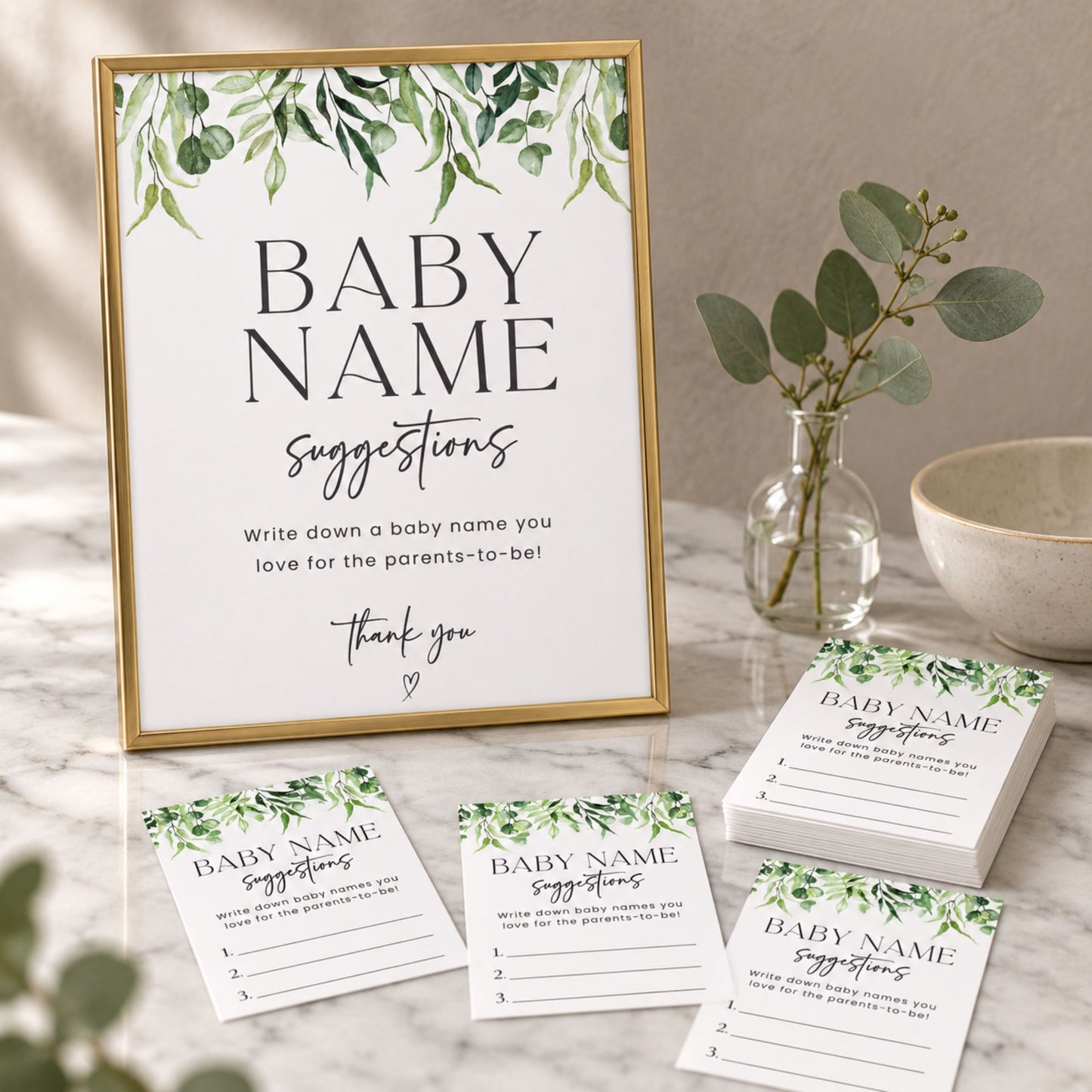 A framed baby name suggestions sign and matching printed cards arranged on a marble surface with a small vase of greenery and a bowl in the background. The cards and sign feature a white base with green leafy border artwork and black text reading 'BABY NAME suggestions' and a thank you message.