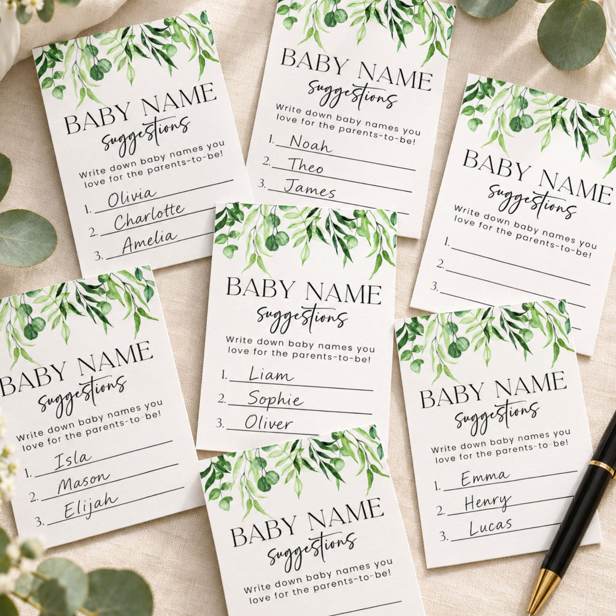 Five baby name suggestion cards with green watercolor foliage at the top, arranged in a flat lay on a beige fabric background with decorative greenery and a black pen.
