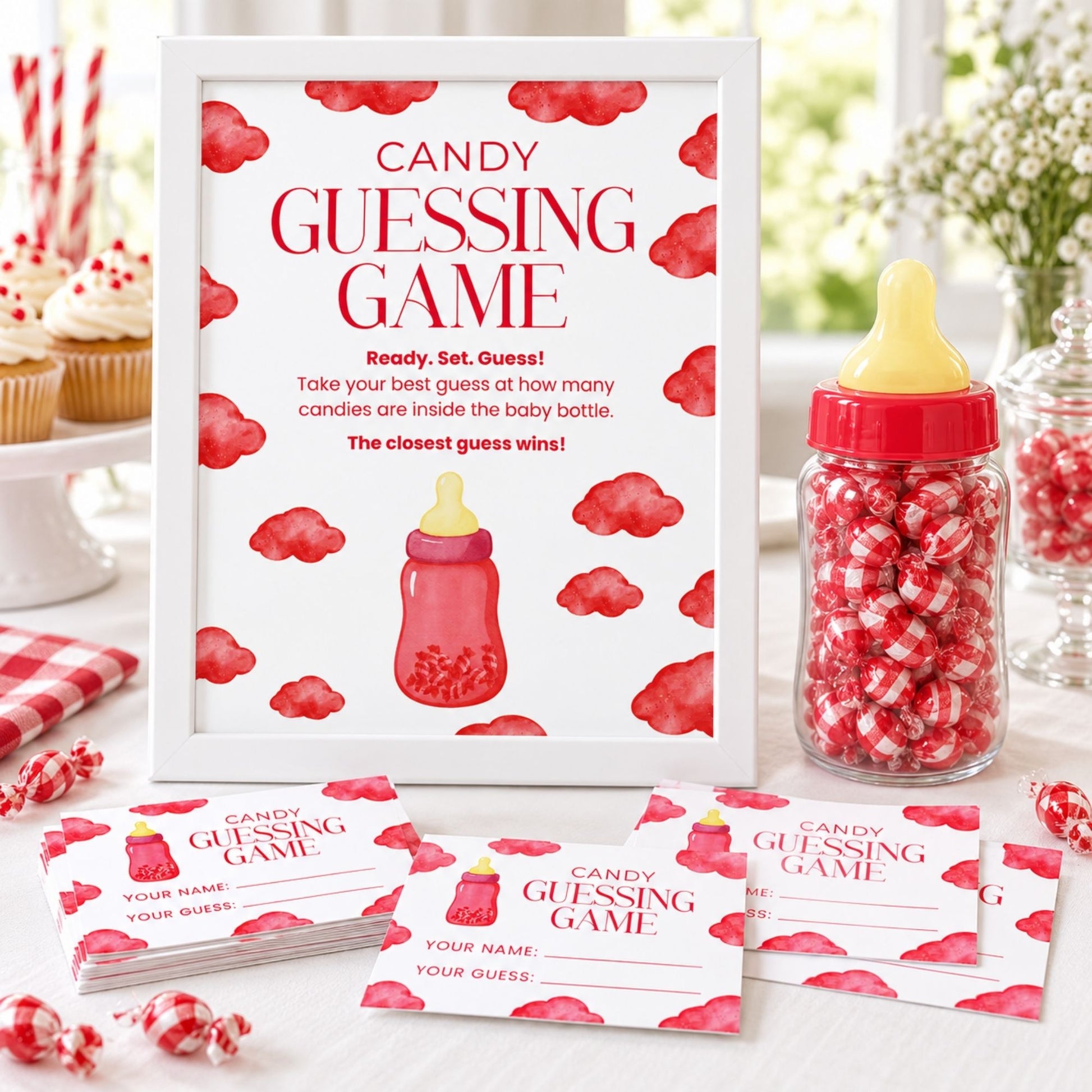 A baby shower candy guessing game set displayed on a table, featuring a large white sign with red watercolor clouds and a baby bottle illustration, matching game cards, a glass jar filled with red-and-white gummy candies, and candy corn in the background.