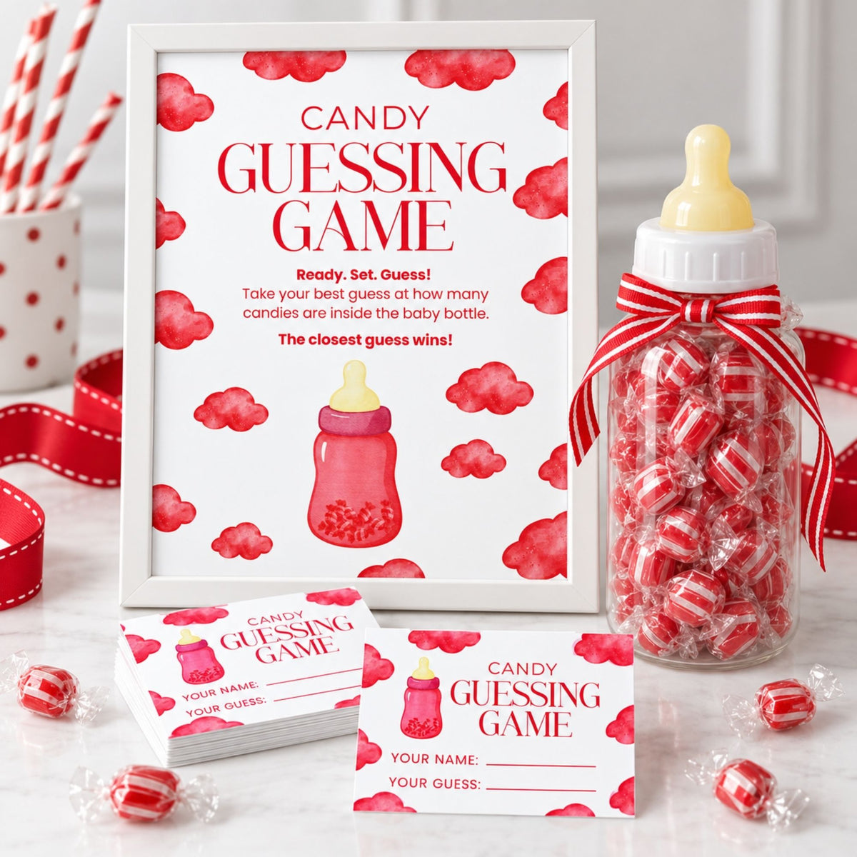 Candy Guessing Game party kit featuring a white framed sign with red watercolor clouds and a baby bottle illustration, a clear bottle filled with red-and-white wrapped candies and tied with a red ribbon, matching name cards, and scattered candy wrappers on a light surface.
