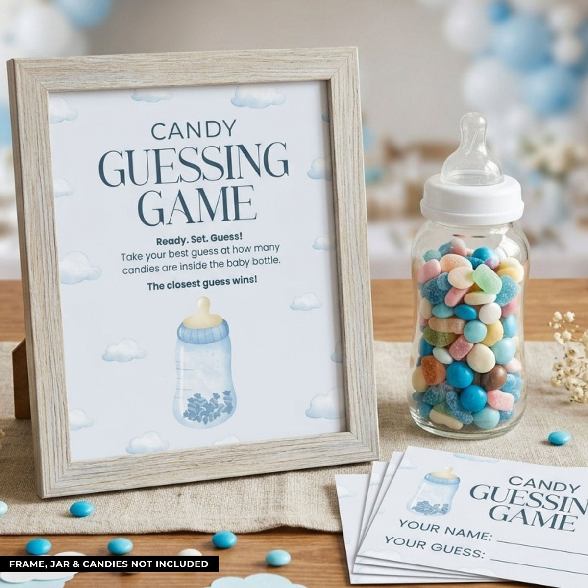 A baby shower candy guessing game setup featuring a framed sign that reads 'CANDY GUESSING GAME' with a baby bottle illustration, a clear baby bottle filled with pastel candies, and printed card inserts on a tabletop.