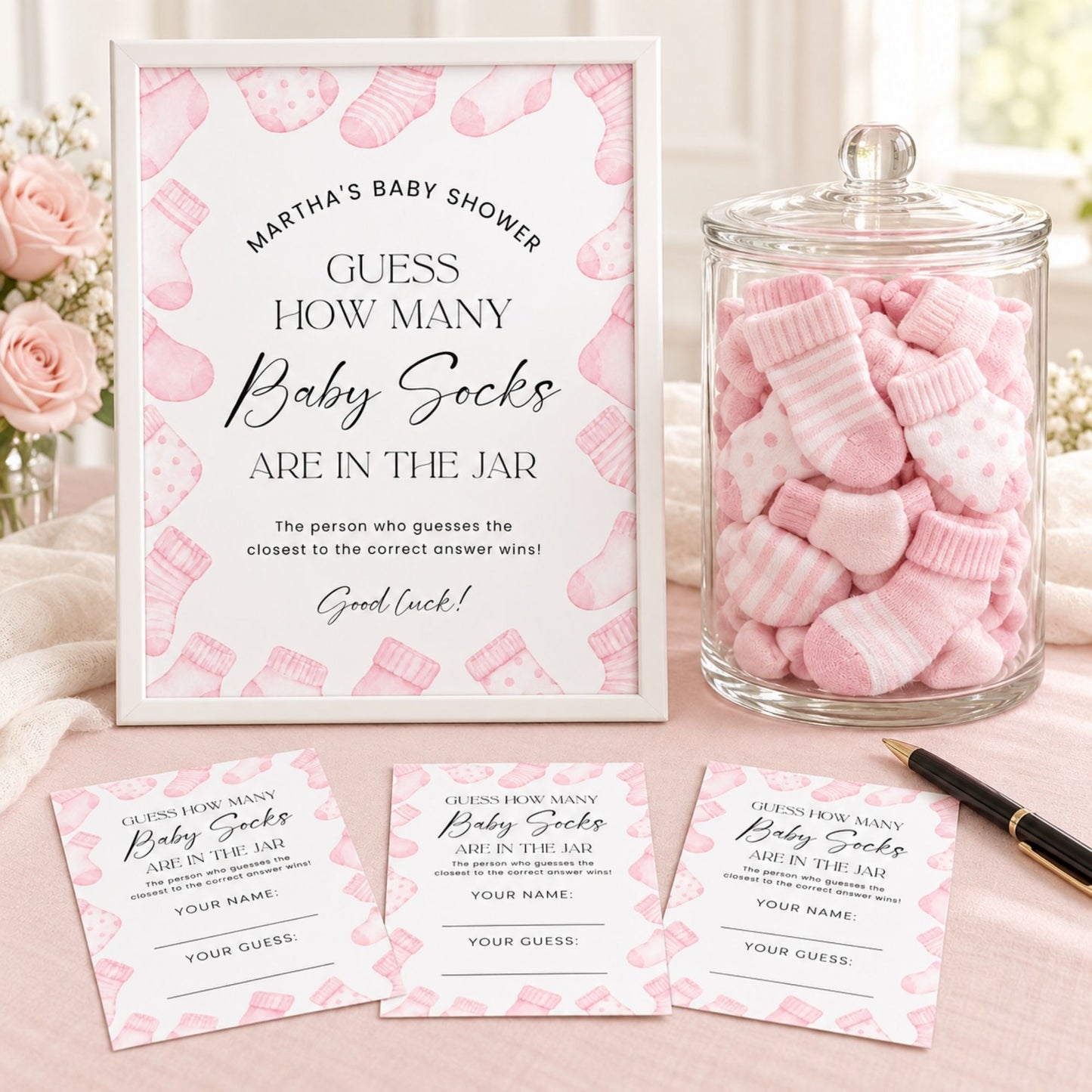 A baby shower game setup featuring a white sign that reads 'Martha's Baby Shower - Guess How Many Baby Socks Are in the Jar,' a clear glass jar filled with pink and white baby socks, and three matching entry cards on a soft pink surface with roses in the background.