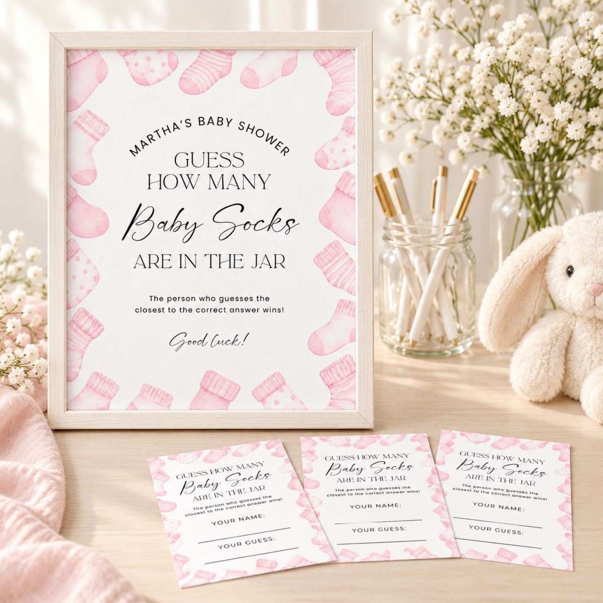 A baby shower game sign and two matching entry cards displayed on a tabletop, with a pink-and-white baby sock pattern, framed sign, flowers, a glass jar with sticks, and a plush bunny in the background.