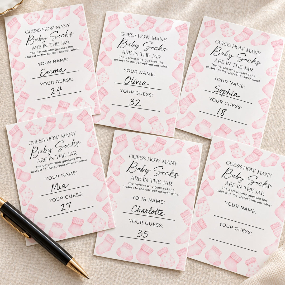 Six pink and white baby socks counting cards arranged on a beige fabric background, with a black and gold pen resting in the bottom-left corner.