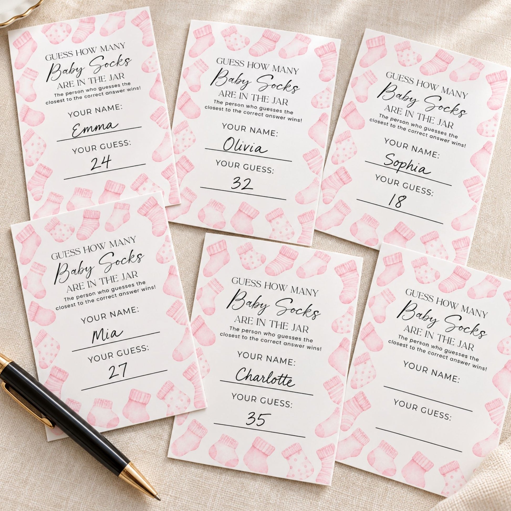 Six pink and white baby socks counting cards arranged on a beige fabric background, with a black and gold pen resting in the bottom-left corner.