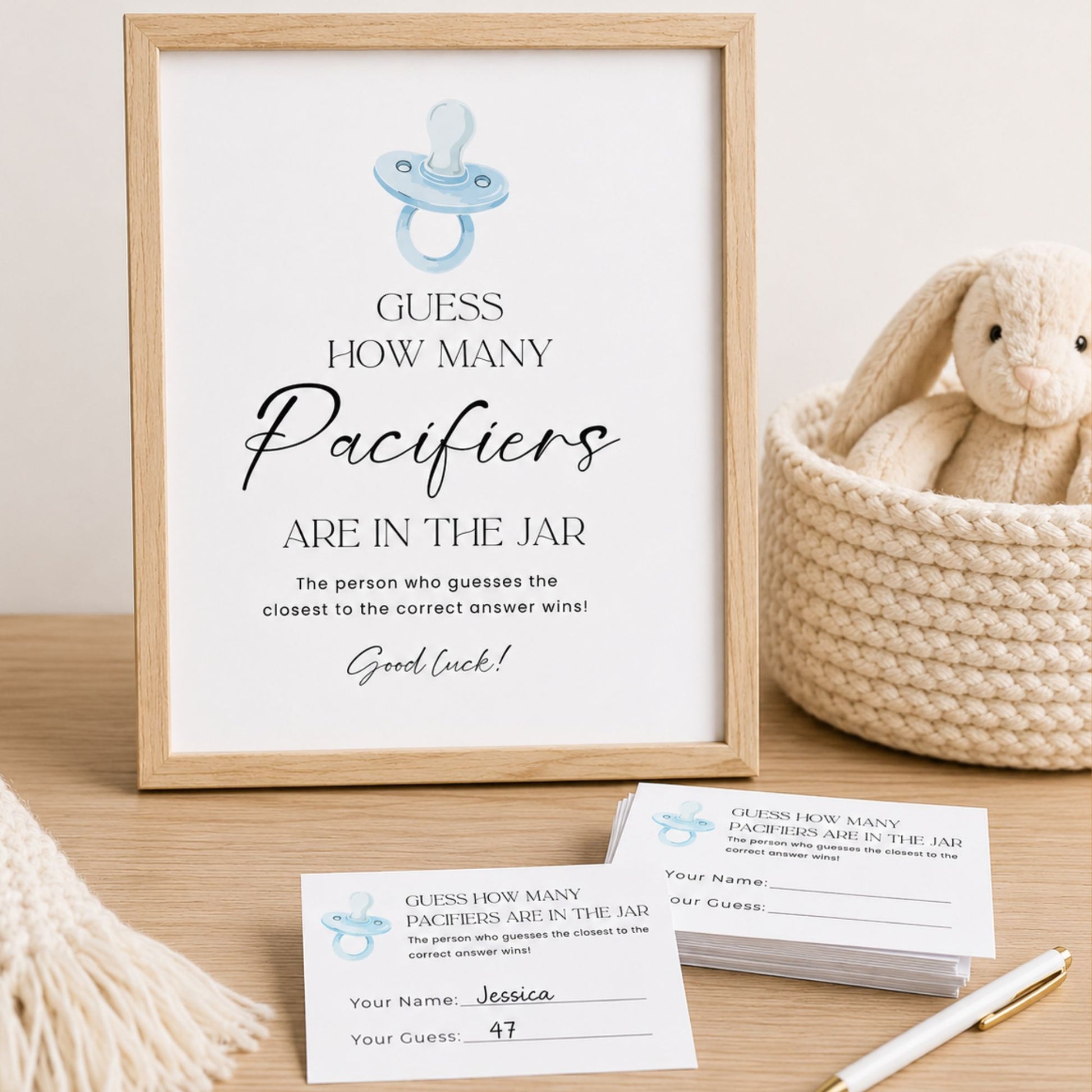 A framed baby shower sign reading 'Guess How Many Paciers Are in the Jar' with a light blue pacifier illustration, displayed on a wooden surface beside a woven basket holding a plush bunny and printed participation cards.