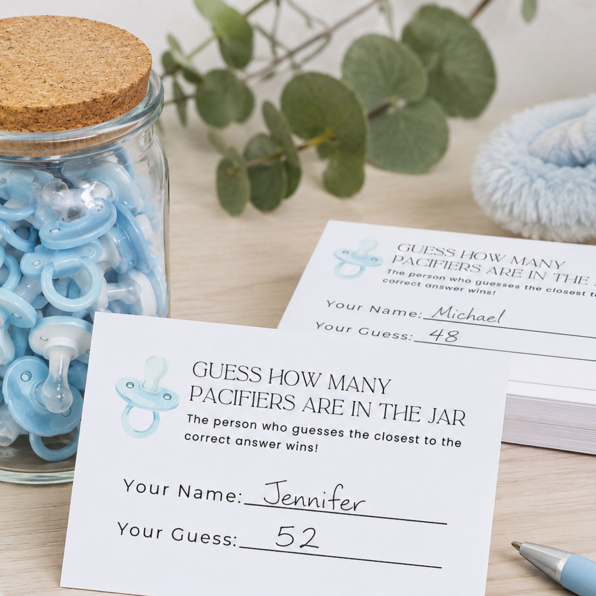 A baby pacifier guessing game card on a light wood tabletop next to a glass jar filled with blue pacifiers, with a eucalyptus branch and a folded blanket in the background.