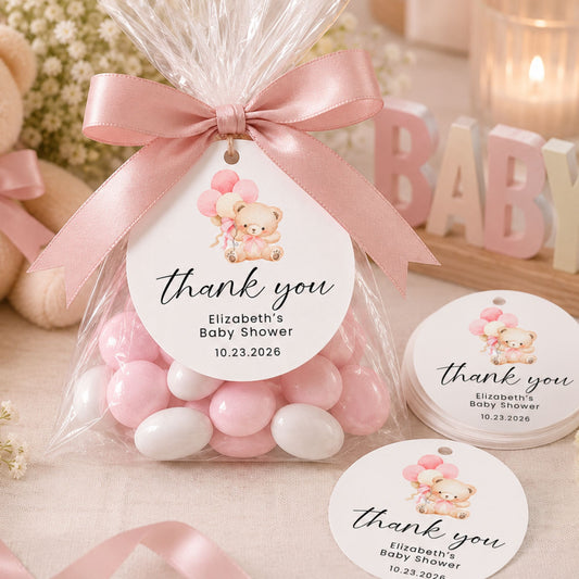 A clear cellophane gift bag tied with a pink satin ribbon, filled with pastel pink and white candy, and topped with a round printed tag that reads 'thank you Elizabeth's Baby Shower 10.23.