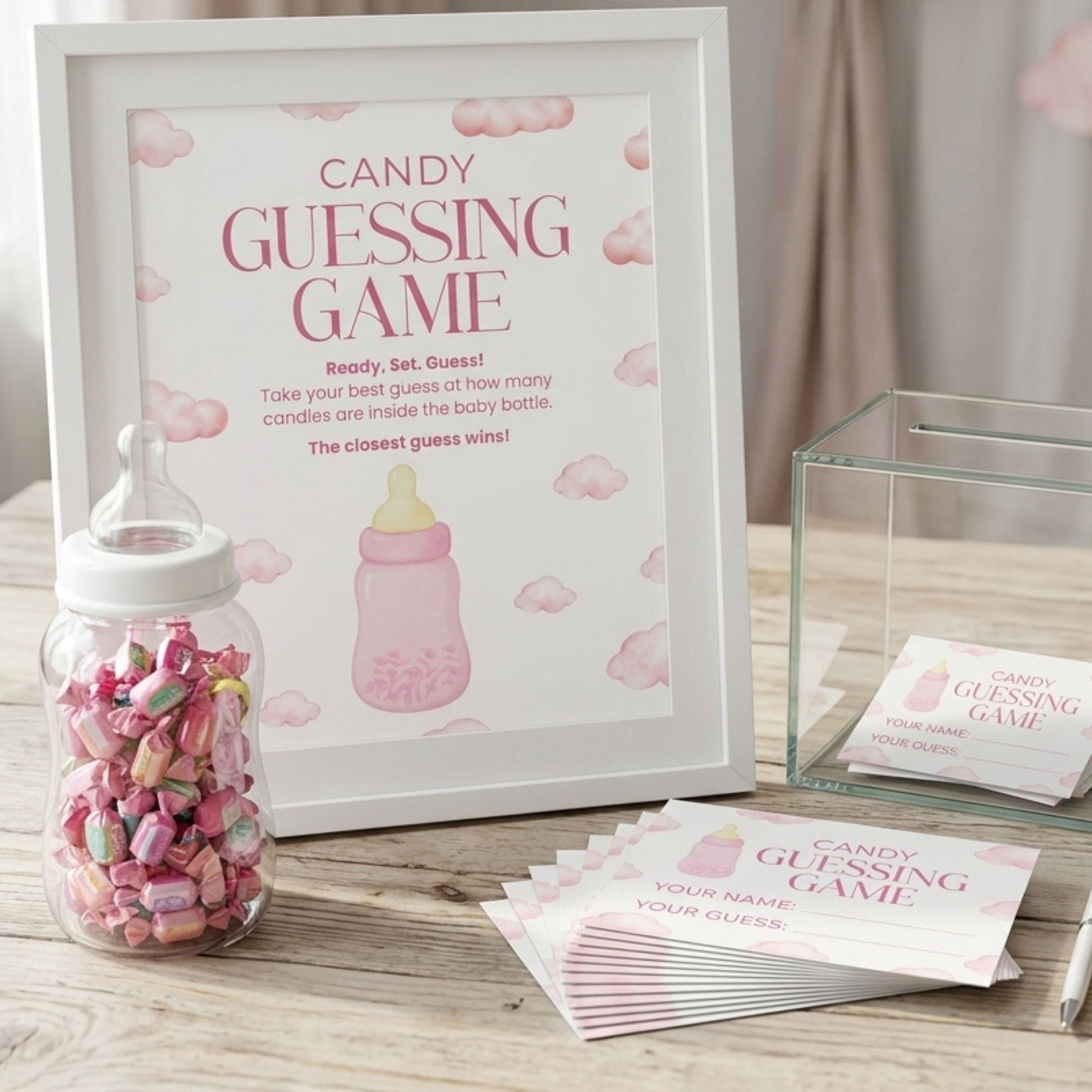 Baby shower candy guessing game kit with a white-framed sign reading 'Candy Guessing Game,' a clear baby bottle filled with pink and multicolored candies, a clear gift box, and printed ticket slips on a wooden surface.