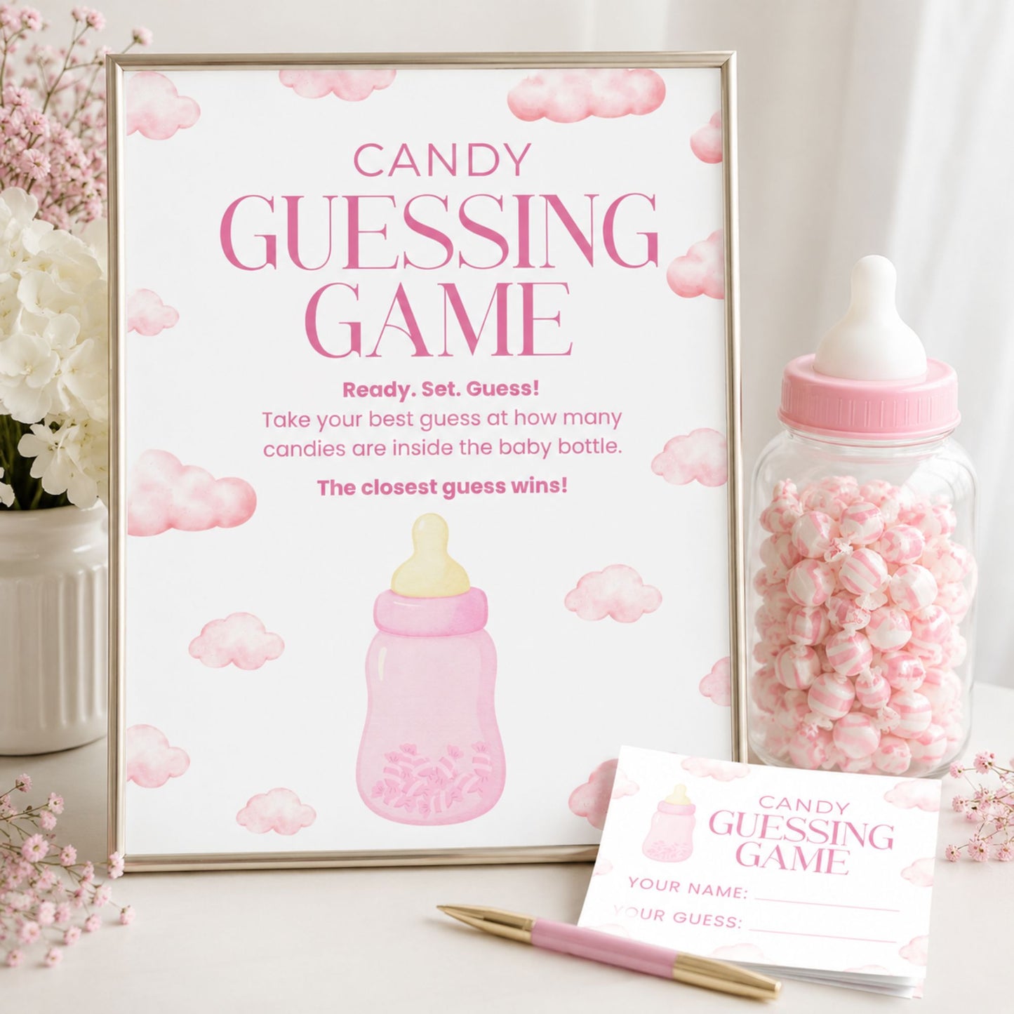A baby shower game setup featuring a framed 'Candy Guessing Game' sign with pink clouds and a baby bottle illustration, a clear bottle filled with pink candy, a matching name card, and a pink-and-gold pen on a white tabletop.