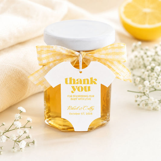 A clear glass jar filled with honey, topped with a white lid and tied with a yellow-and-white gingham ribbon. A white baby onesie-shaped tag hangs from the ribbon, printed with the words 'thank you' and baby shower gift message text.