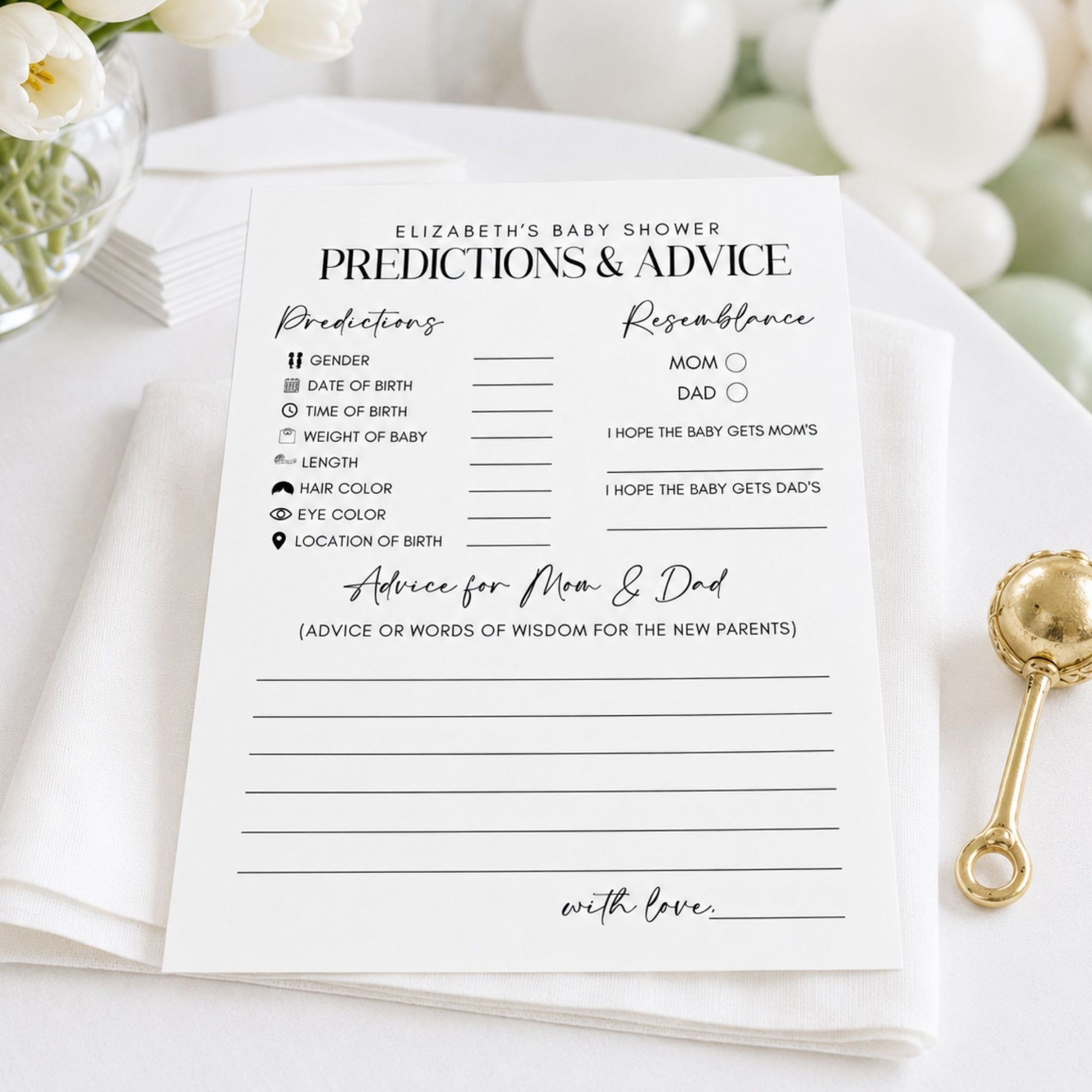 A baby shower prediction card laid on a white surface with a gold baby rattle nearby and soft white flowers in the background.