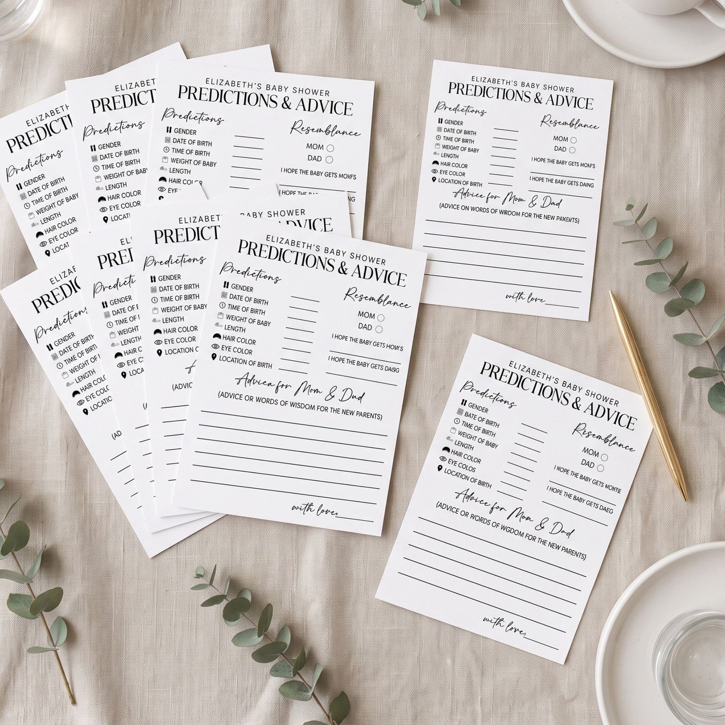 A styled flat-lay of several printed prediction and advice cards for a baby shower, arranged on a light wood surface with eucalyptus leaves, a gold pen, and white dishes.