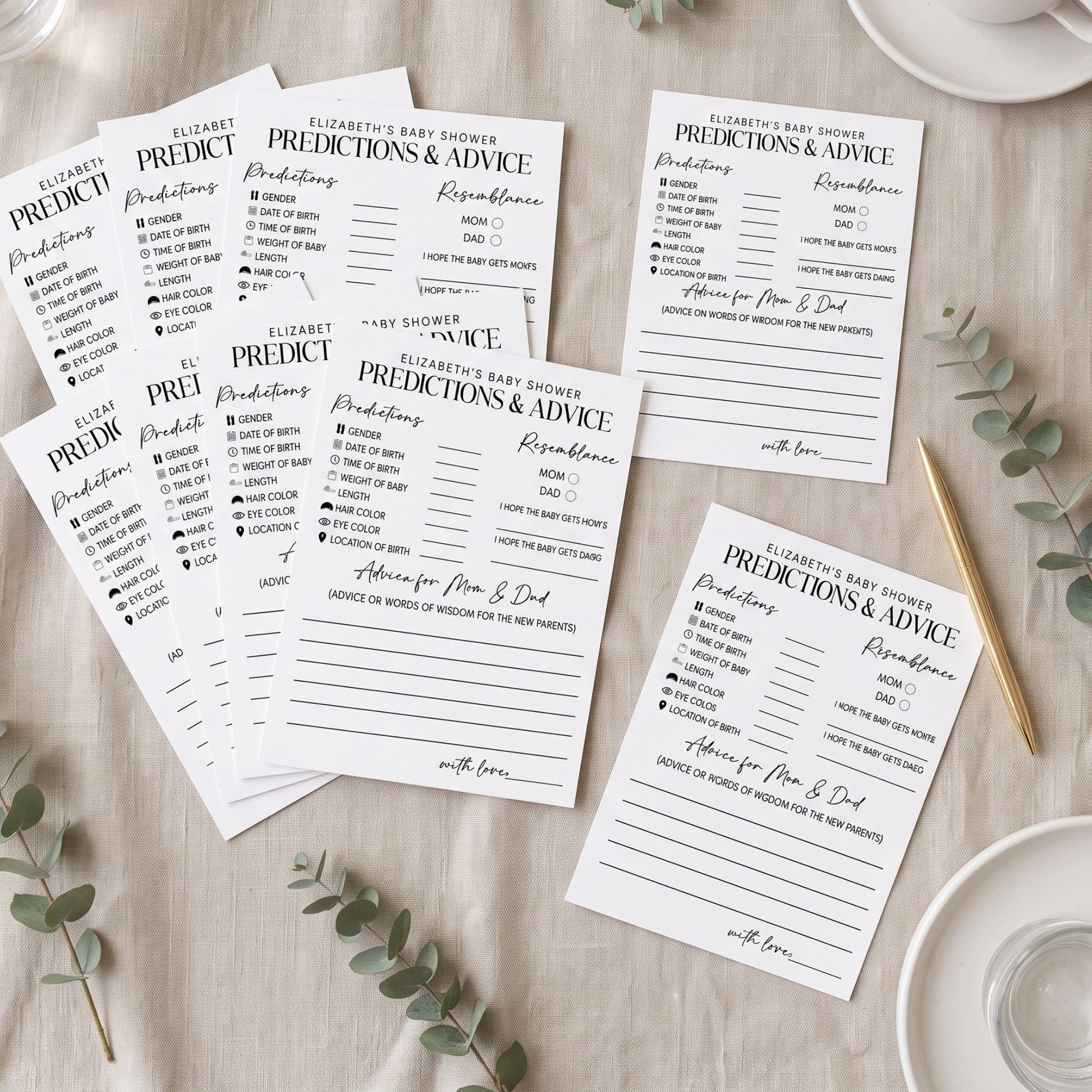A styled flat-lay of several printed prediction and advice cards for a baby shower, arranged on a light wood surface with eucalyptus leaves, a gold pen, and white dishes.
