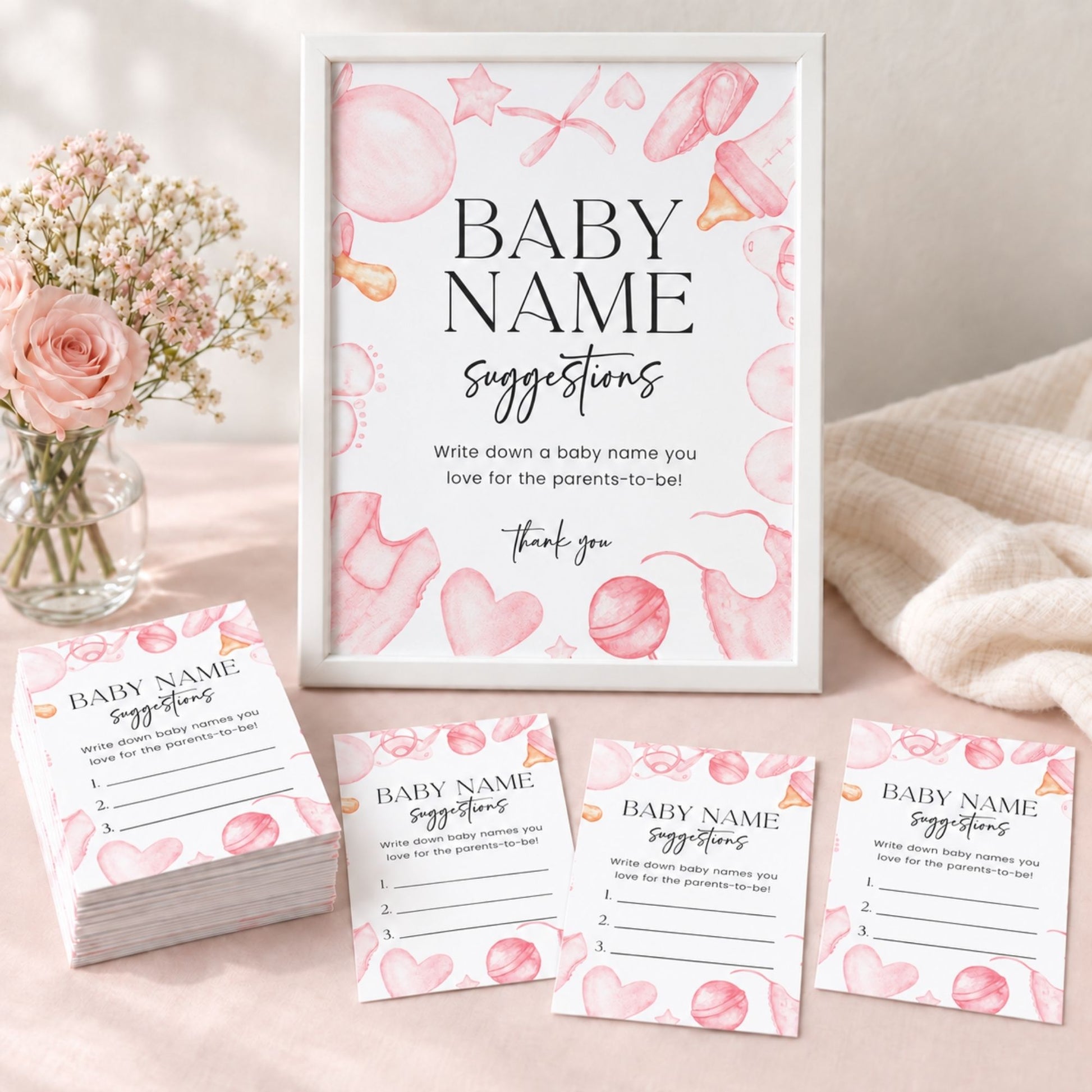A baby shower game set featuring a white-framed sign and matching cards with pink watercolor hearts, balloons, and balls, styled on a soft pink surface with flowers and a cream throw blanket.