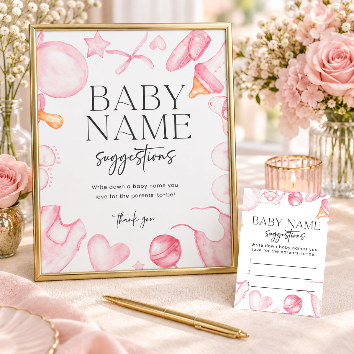 A baby name suggestion sign and matching suggestion card styled on a table with pink flowers, a gold pen, and a gold candlestick. The sign features watercolor baby-themed illustrations including hearts, stars, and pacifiers in pink and peach tones, with black text reading 'BABY NAME suggestions'.