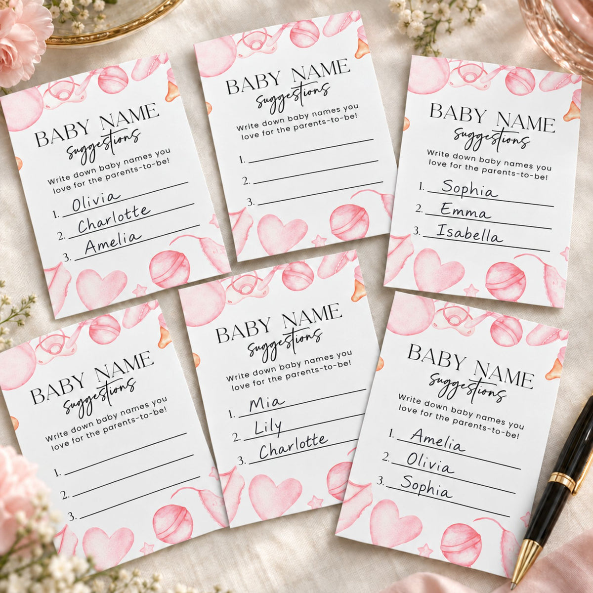 Six printed baby name suggestion cards arranged on a light fabric background with pink flowers and a black pen. The cards are white with pink watercolor hearts and the title 'BABY NAME suggestions' in black text.