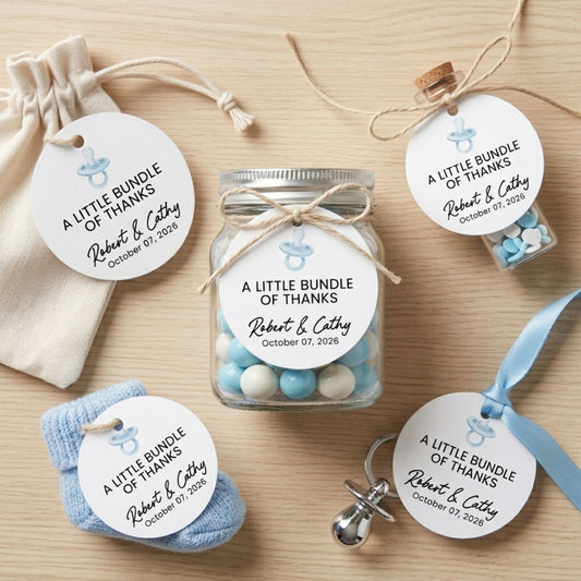 Personalized baby keepsake gift set on a light wood background, including a small jar of blue and white candies, a cream drawstring pouch, a blue sock, a pacifier, and round paper tags reading 'A Little Bundle of Thanks Robert & Cathy October 01, 2026' with a baby silhouette.