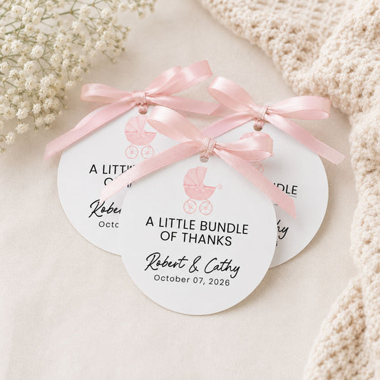 Three round white baby shower favor tags tied with light pink ribbons, decorated with a small pink baby carriage graphic and personalized text, arranged on a soft cream fabric with dried flowers and a knitted blanket.