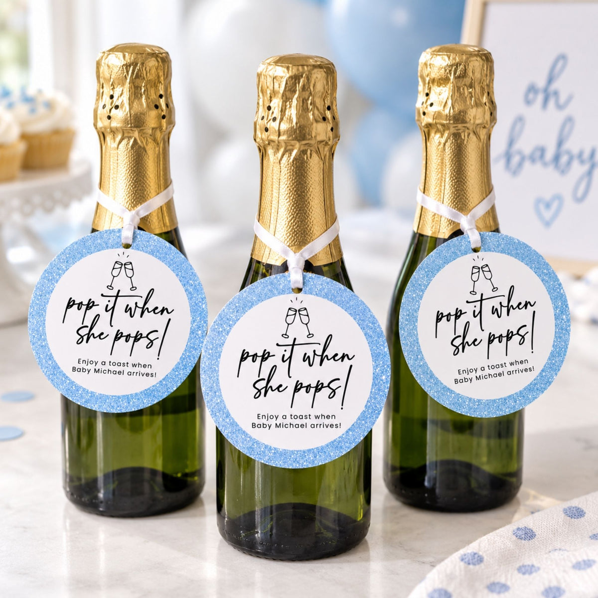 Three mini champagne bottles with gold foil neck wraps and round blue glitter tags that read 'pop it when she pops! Enjoy a toast when baby Michael arrives!' are arranged on a table in a decorated party setting.