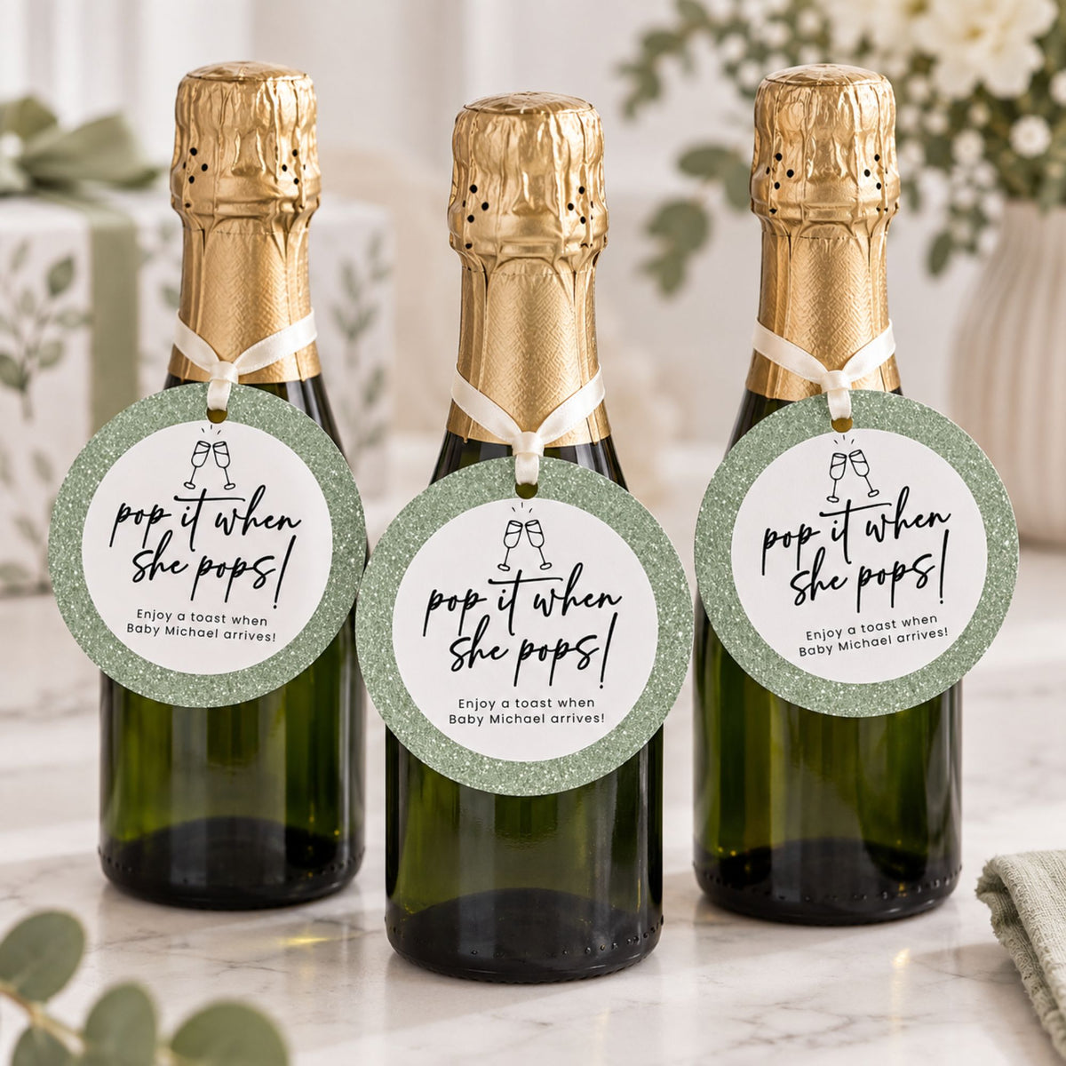 Three mini champagne bottles with gold foil tops and round green glitter hang tags that read 'pop it when she pops! Enjoy a toast when baby Michael arrives!' on a light marble surface with soft floral decor in the background.