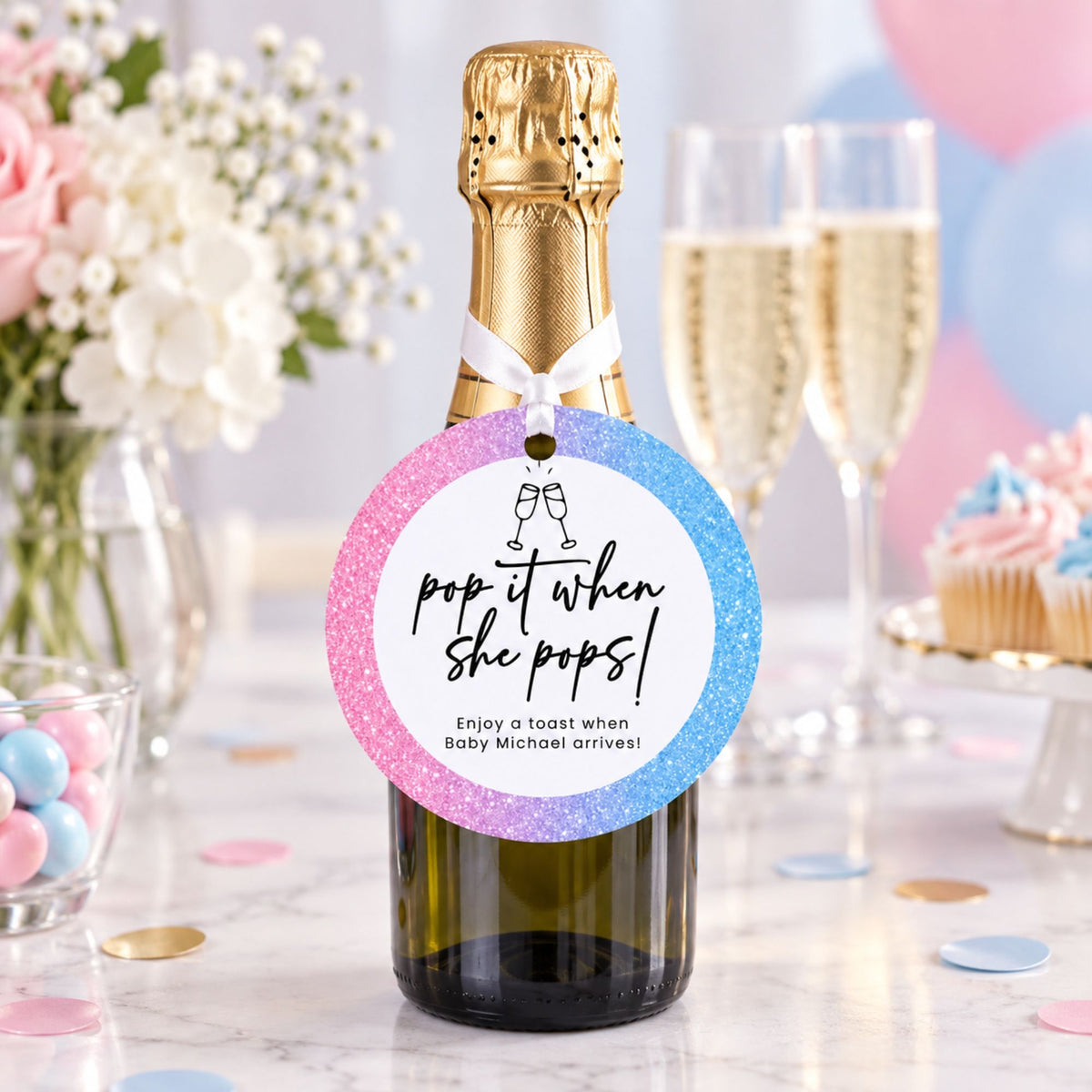 A personalized champagne bottle with a gold foil top and a round glitter tag that reads 'pop it when she pops! Enjoy a toast when Baby Michael arrives!' The bottle is styled on a marble table with champagne flutes, flowers, and cupcakes in the background.