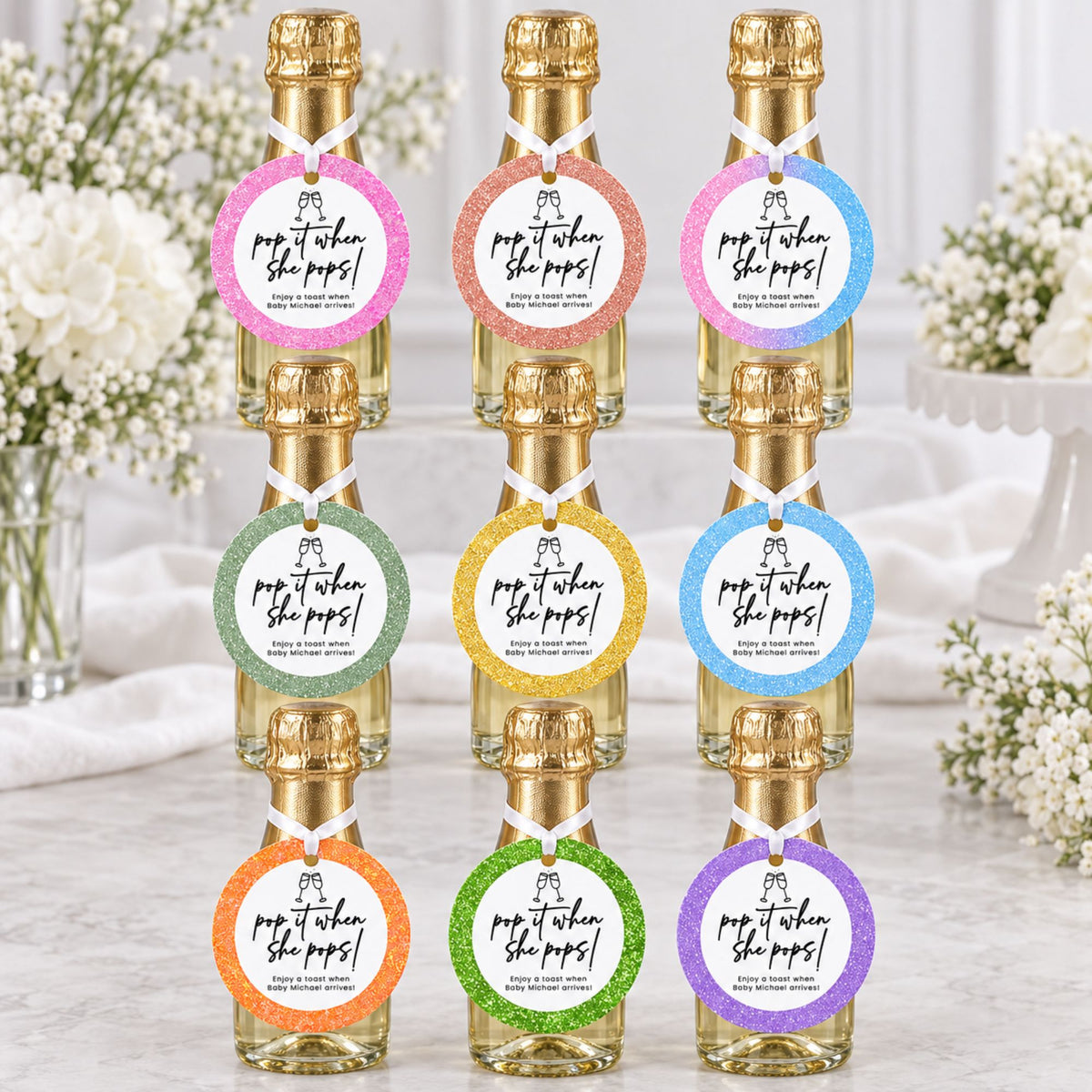 Eight mini champagne bottles arranged in two columns on a light marble surface, each with a gold foil top and a round glittery paper label that reads 'pop when she pops!'. The labels come in pink, orange, green, yellow, blue, and purple colorways, with white flowers blurred in the background.